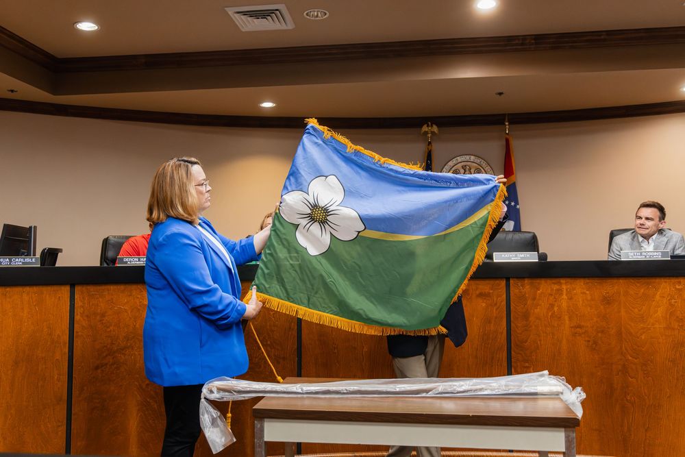 Woman in blue blazer displays green flag with white flower emblem at a government council meeting.