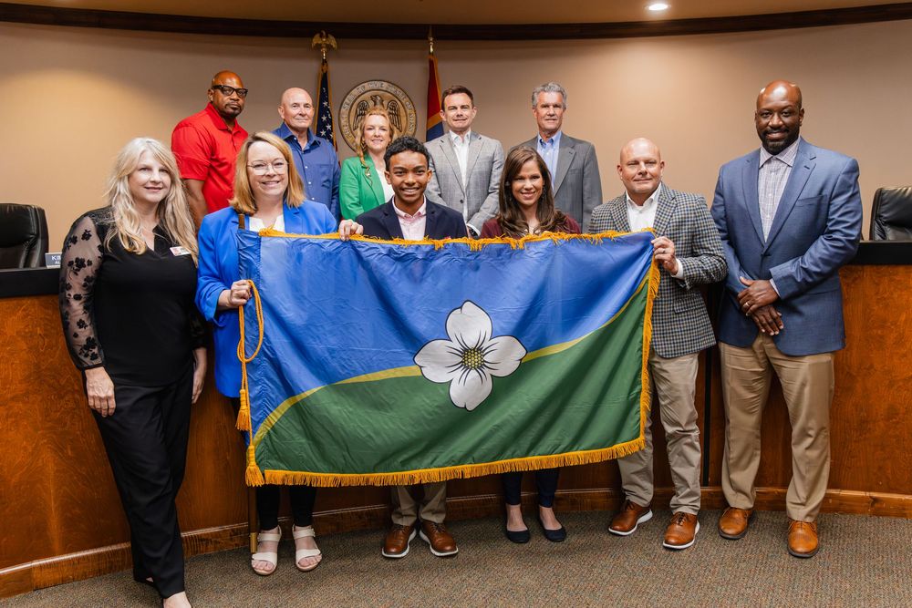 Group of officials proudly holding a blue and green flag with a white magnolia flower in a government meeting room.