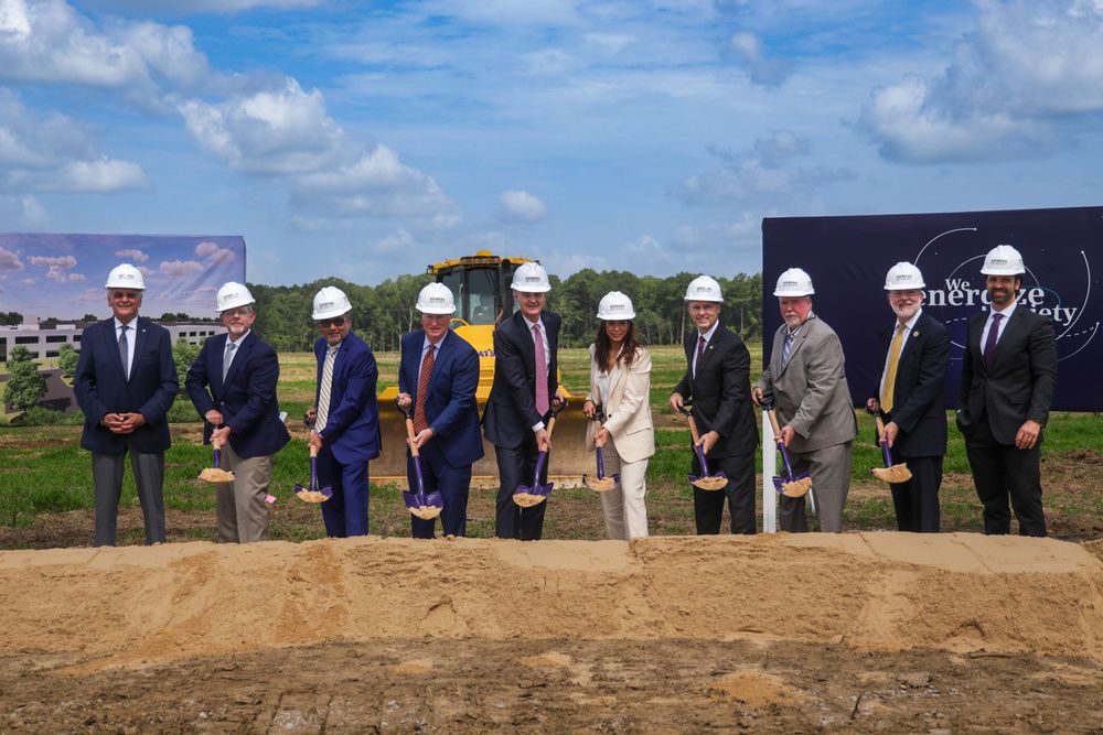 Group of executives in white hard hats holding shovels at a corporate groundbreaking ceremony outdoors.