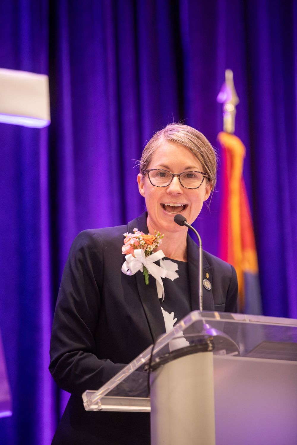 Woman in black blazer with floral corsage speaking at a glass podium before a purple curtain backdrop.