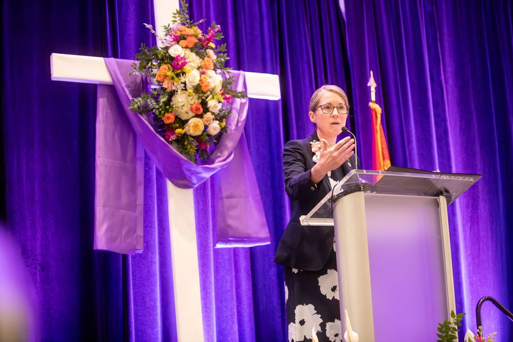 Woman speaking at podium near floral cross arrangement with purple curtain backdrop at religious event.