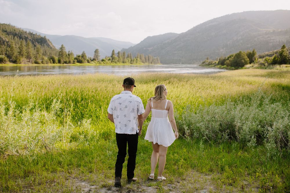 Couple holds hands while walking through a scenic mountain meadow with a lake in the background.