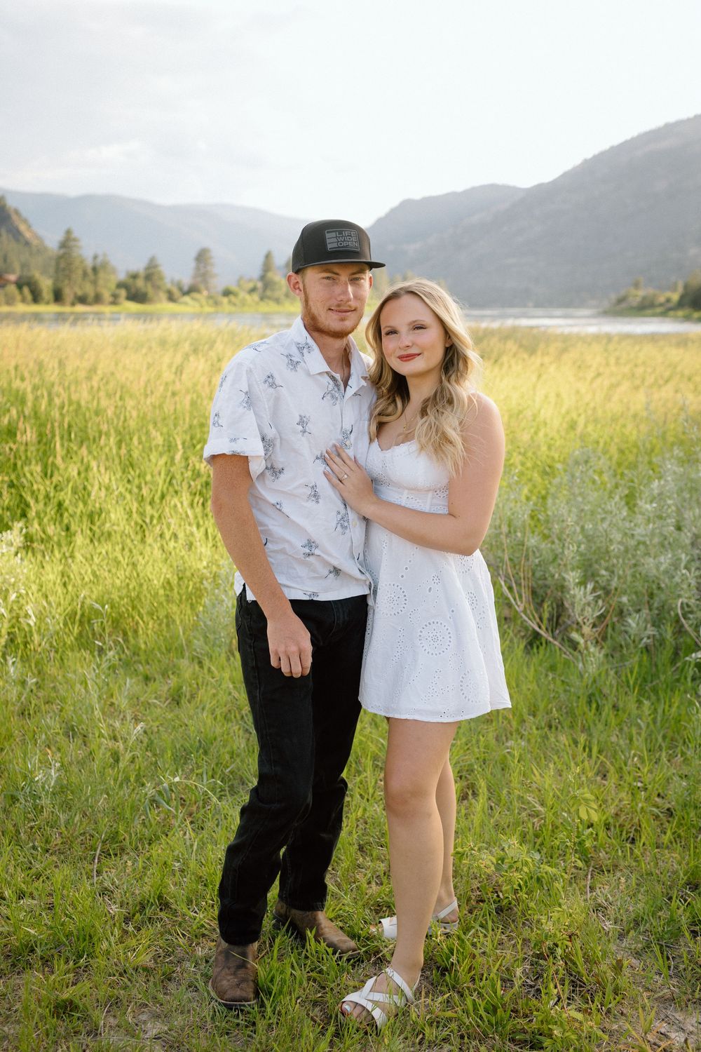 Couple embraces in a sunny meadow with mountains and lake in the background.