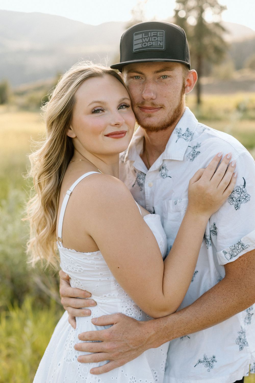 Romantic couple embraces in a sunlit field with mountains in the background.