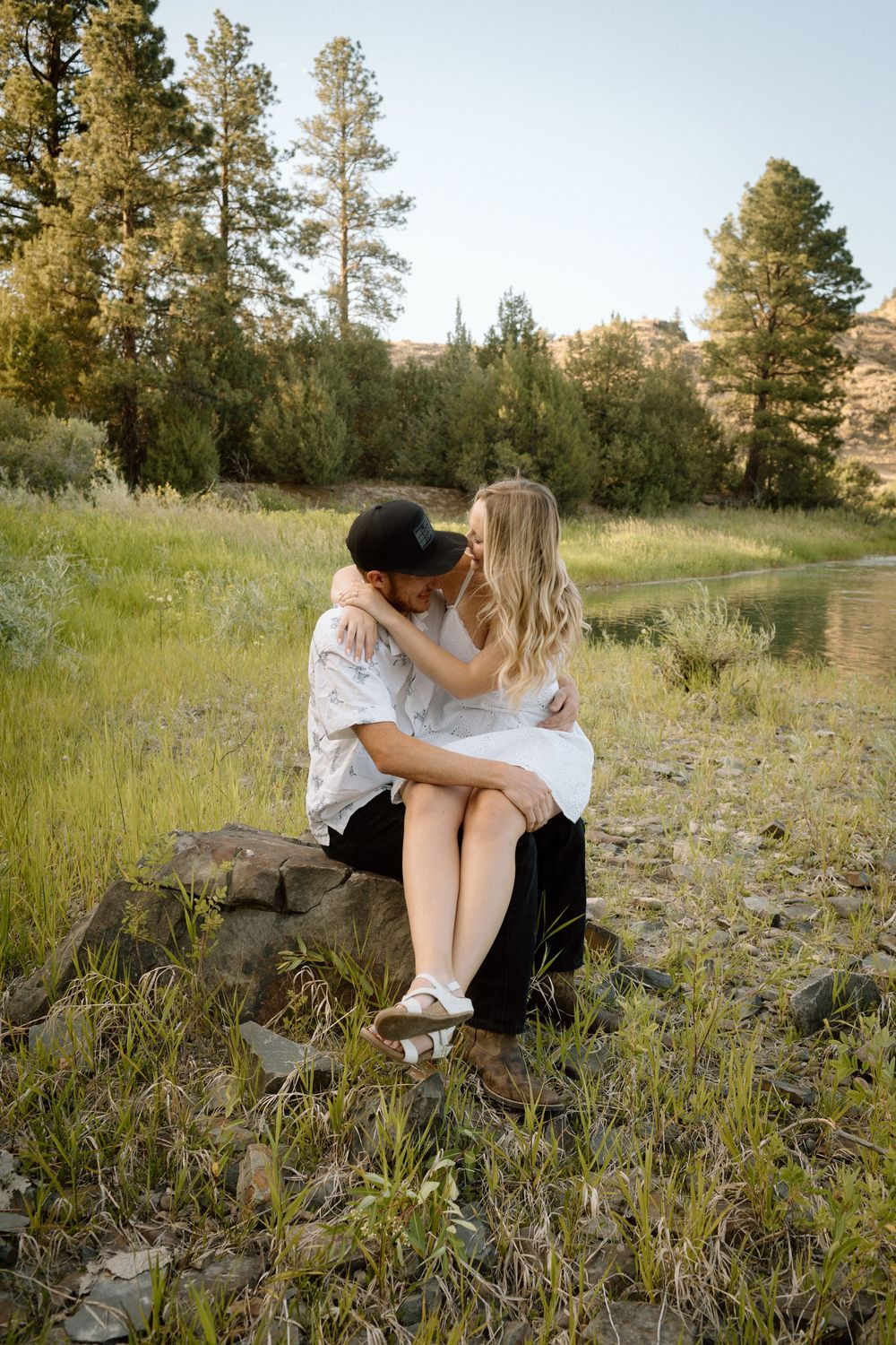 Couple shares tender moment among pine trees with mountain lake backdrop.