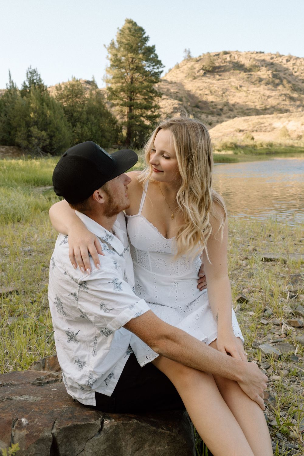 Couple sits intimately on rocky shore with mountain lake in background.