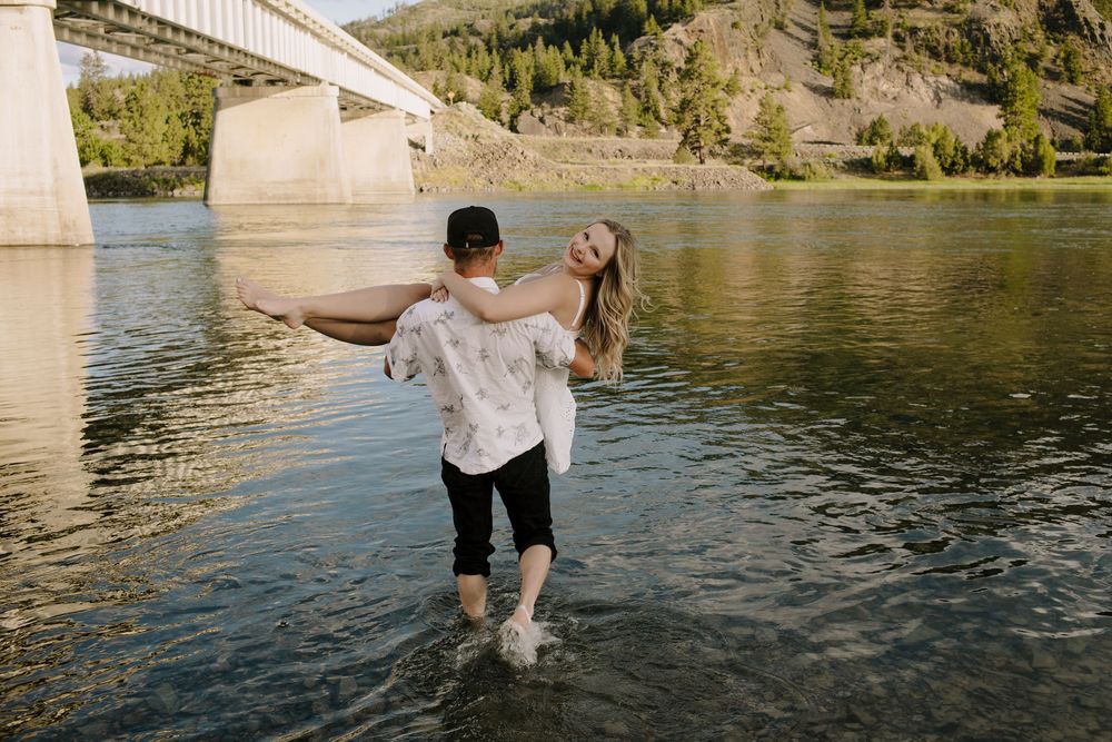 Playful couple wades in mountain lake water during outdoor photo session.