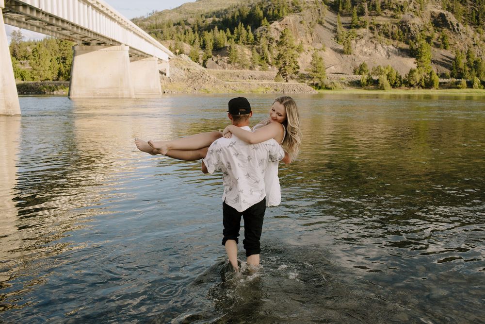 Couple splashes and plays in shallow lake water with mountains in background.