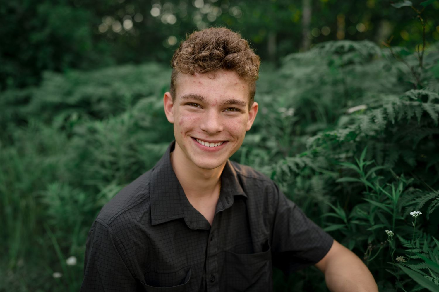 A happy person in a black shirt sits outdoors surrounded by lush green foliage on a sunny day.