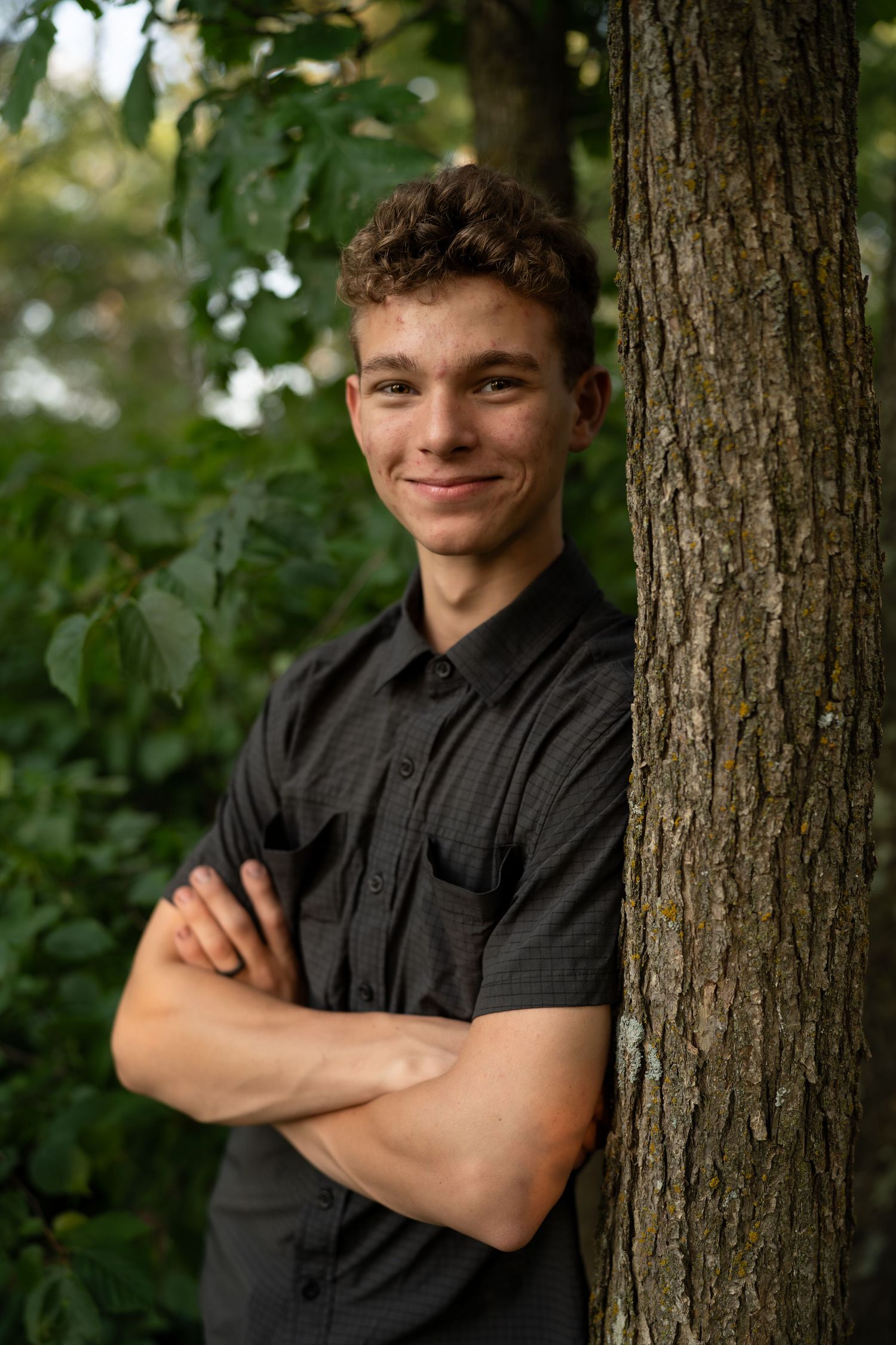 A young person in a black button-up shirt poses against a tree trunk in a series of casual outdoor portraits.