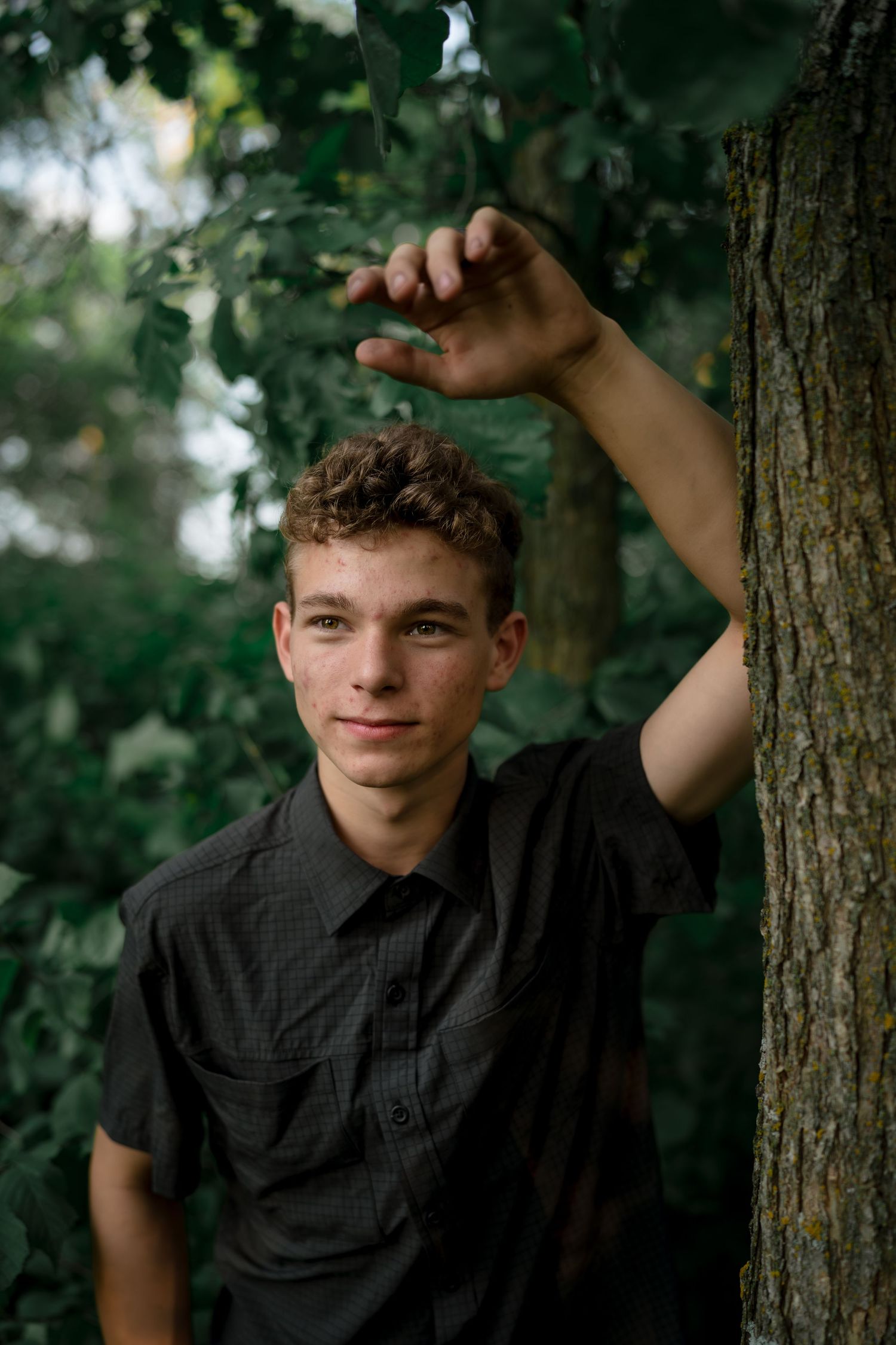 A young person in a black shirt leans against a tree trunk in a natural outdoor setting with green foliage.