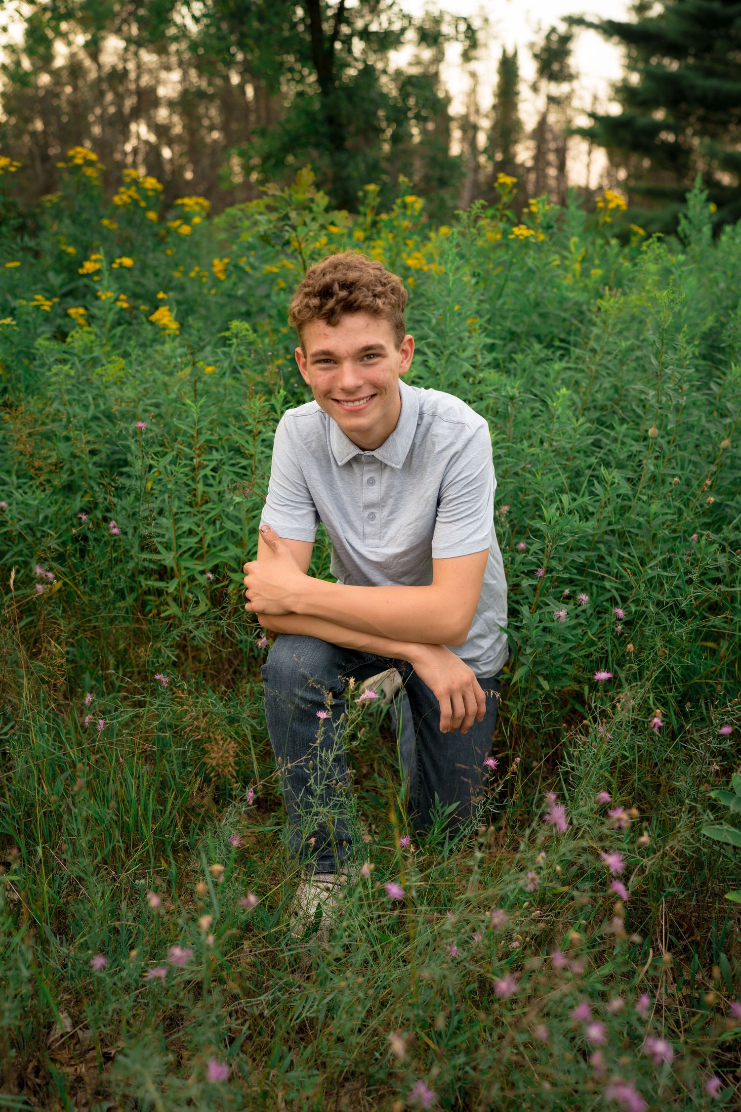 A person sits surrounded by tall wildflowers and greenery in a natural outdoor setting during golden hour.