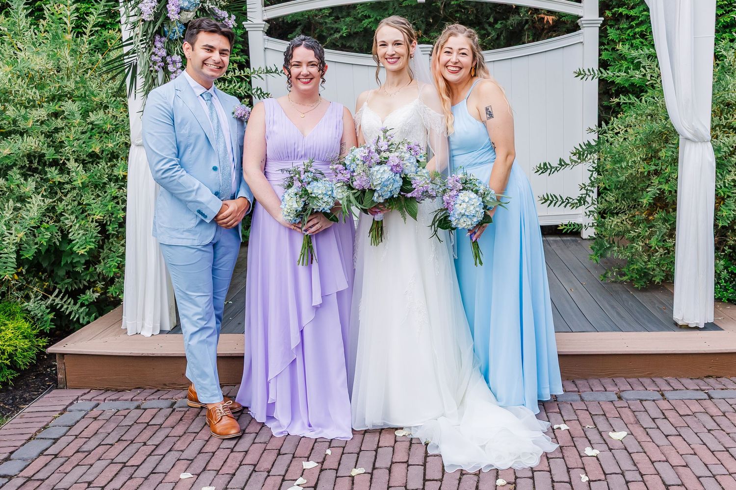 Wedding party members in coordinating pastel outfits pose together on a brick patio in front of a white gazebo.