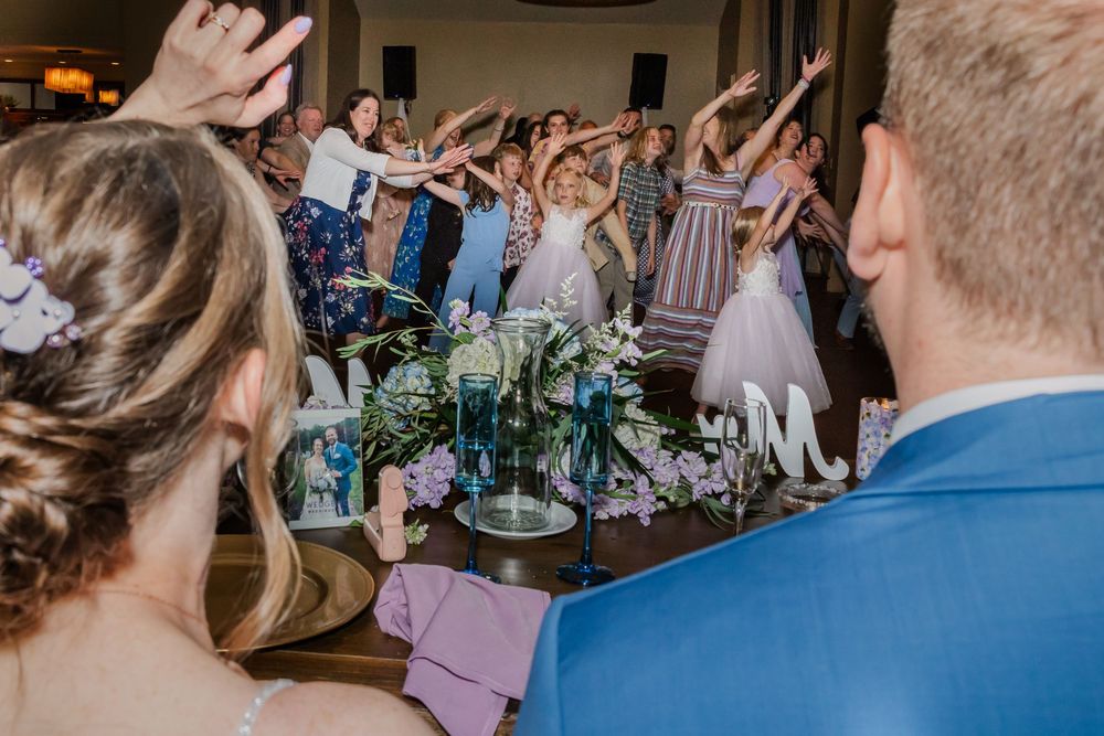 Wedding reception guests raise their hands in celebration as floral centerpieces adorn the tables at an indoor venue.