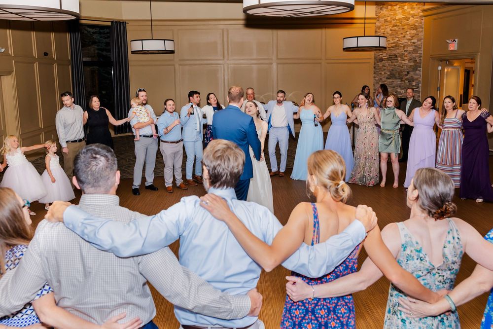Wedding guests form a circle around the dance floor during a reception while wearing blue and purple attire.