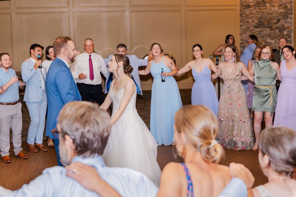 Wedding guests form a line dance circle in a rustic reception venue with exposed brick walls and wooden floors.
