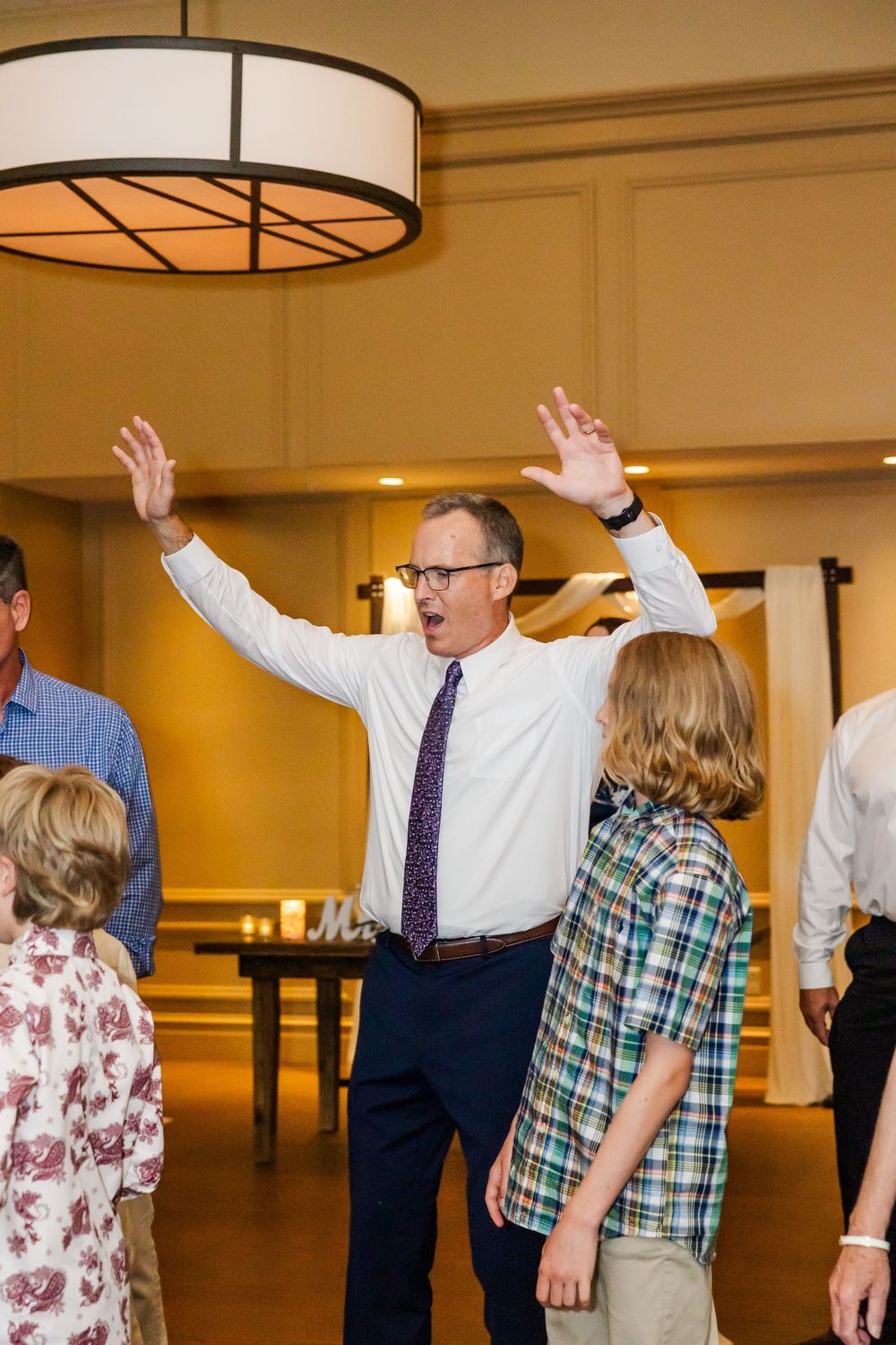 Someone dances with enthusiasm at an indoor event, raising both arms in the air while wearing a white shirt and purple tie.