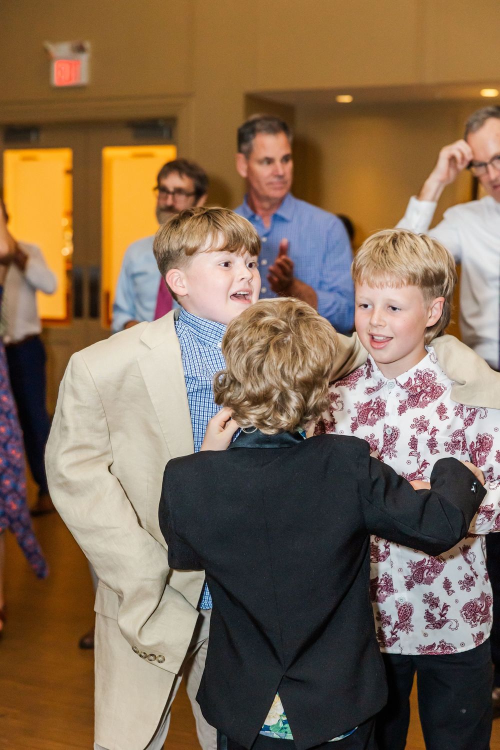 Young boys in formal attire interact and laugh together at an indoor social gathering with adults in background.
