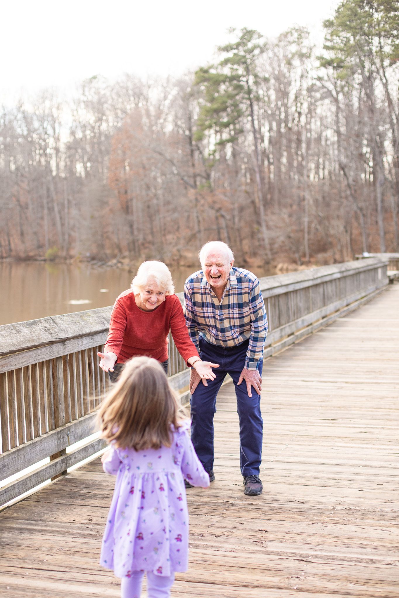 a granddaughter runs to grandparents on a wooden boardwalk over a lake, with trees visible in the background at Yates Mill