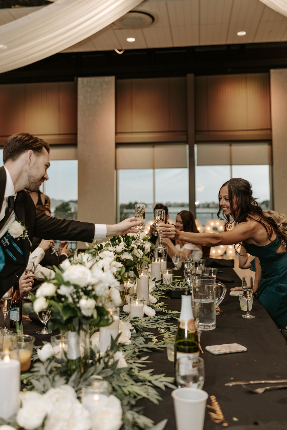Wedding guests toast with champagne glasses across an elegantly decorated reception table with white floral centerpieces.