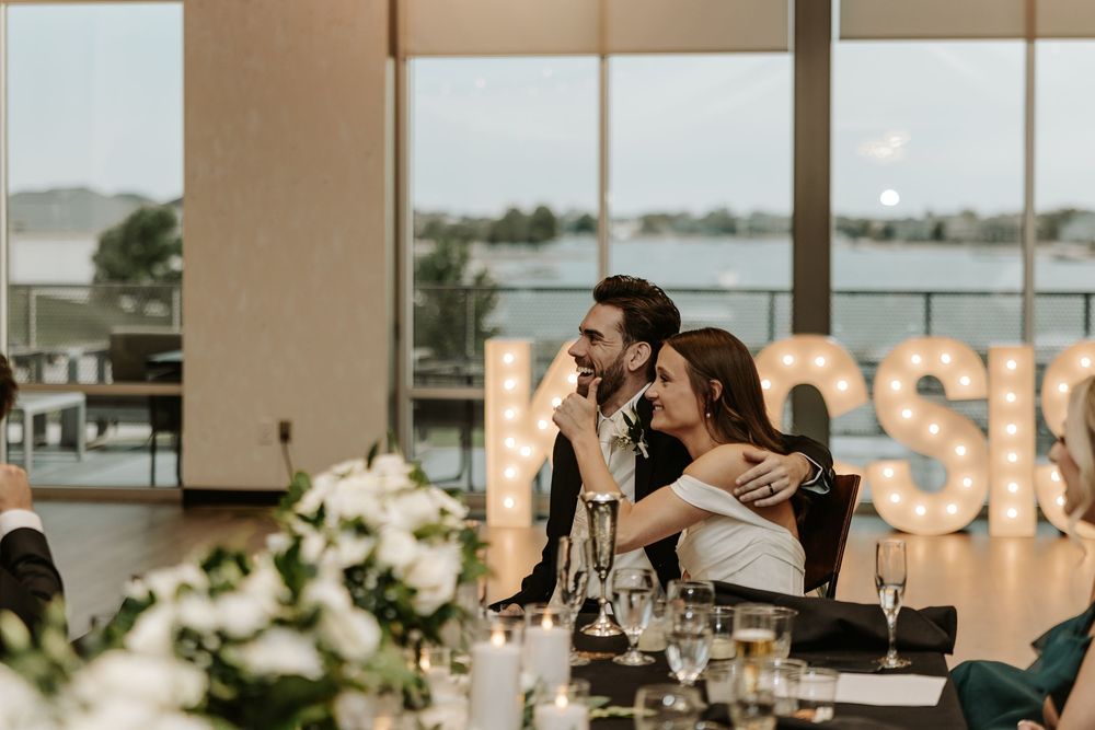 A couple embraces at a waterfront reception venue with illuminated letters and floral decor on the table.
