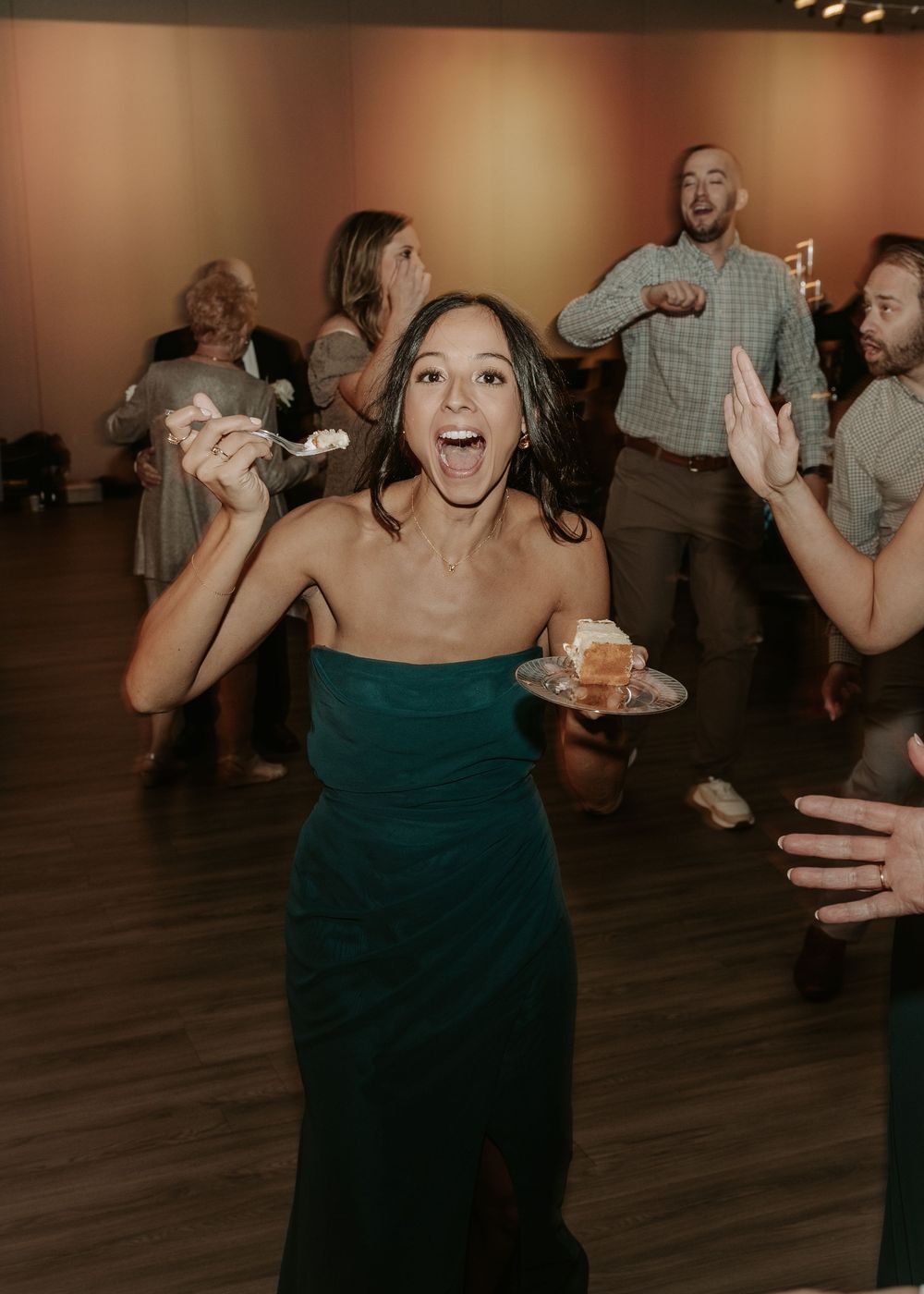Wedding guest in emerald green dress enthusiastically dances with dessert at reception celebration.
