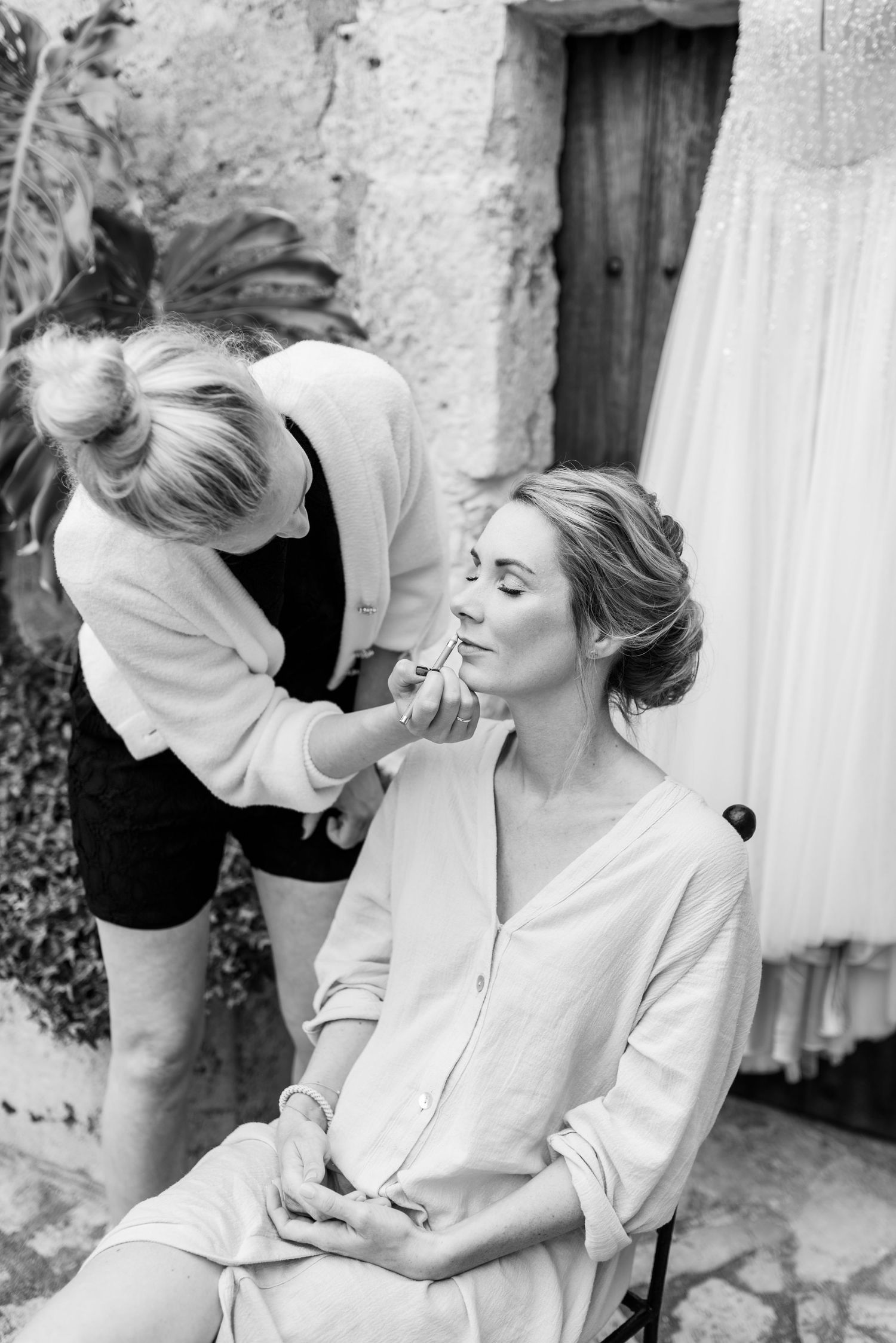 Schwarz-Weiß-Fotografie einer Visagistin, die einer Braut Make-up aufträgt an ihrem Hochzeitsmorgen. Das Brautkleid hängt im Hintergrund.