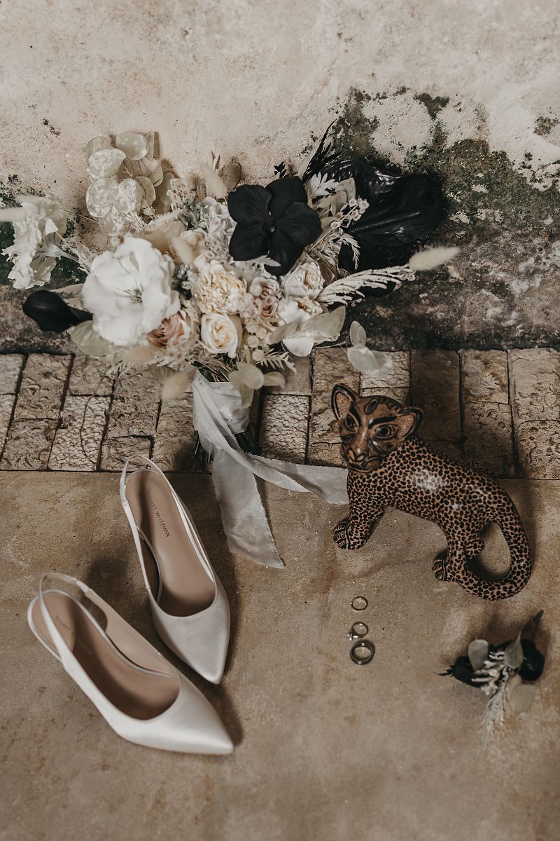 Wedding flat lay with bridal bouquet, ivory heels, rings, boutonniere, and leopard figurine on stone floor.