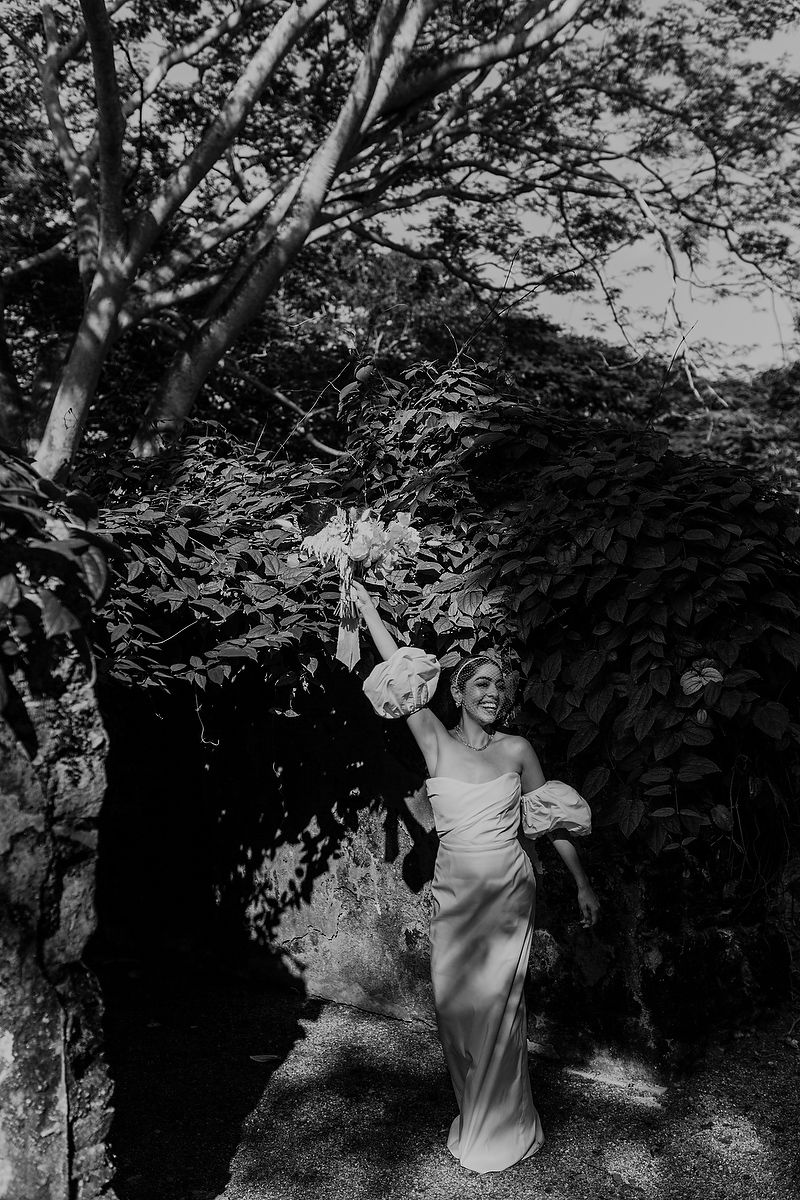 Joyful bride in white gown raising bouquet triumphantly under lush green garden archway.