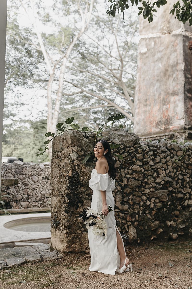Bride in off-shoulder white gown holding bouquet poses by rustic stone wall surrounded by lush greenery.