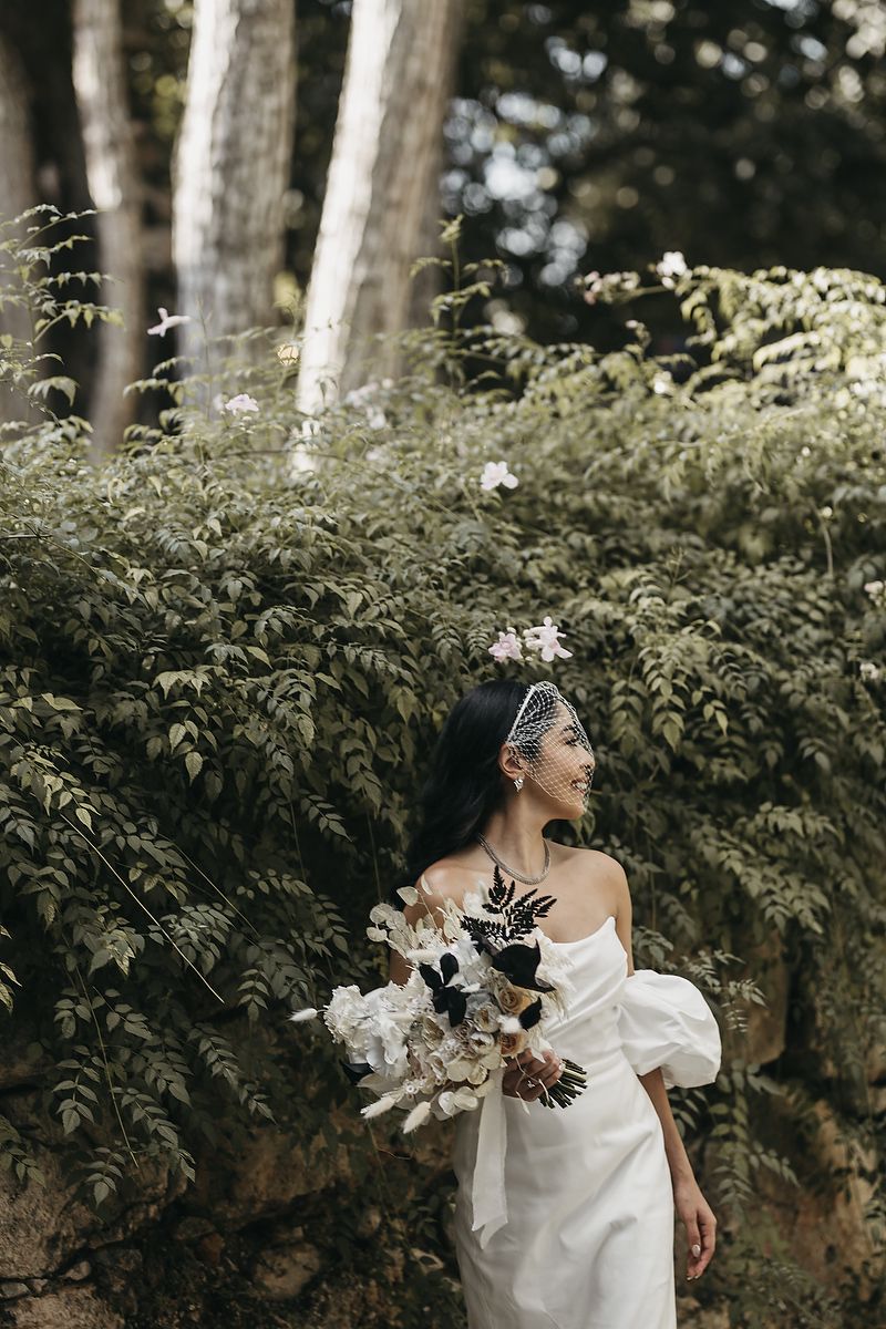 Bride in white off-shoulder gown holds black and white bouquet, standing among lush green foliage in a forest setting.