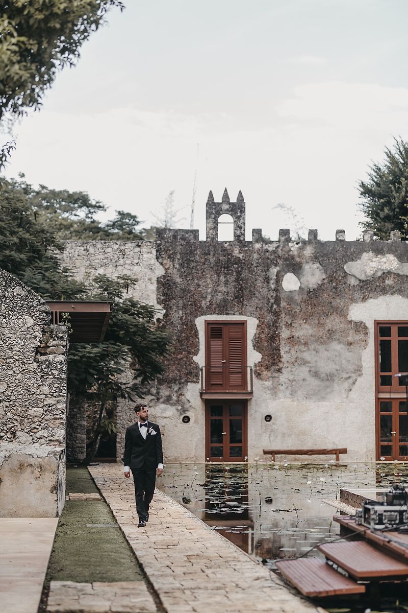 Groom in black tuxedo walking along stone pathway at rustic historic venue with ivy-covered walls and bell tower.