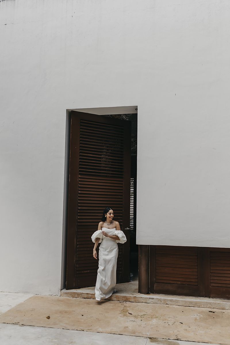 Woman in white off-shoulder gown standing in a doorway with wooden shutters against a white wall.