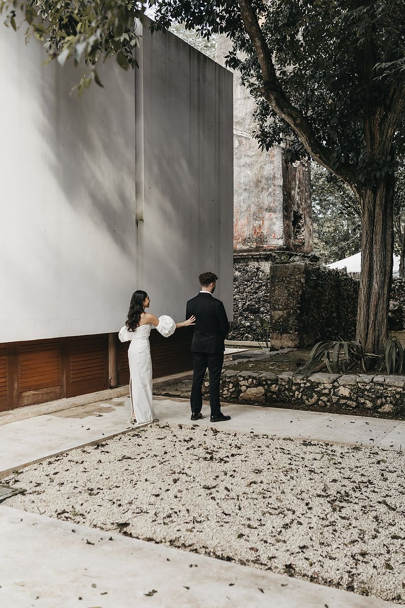 Bride in white gown and groom in black tuxedo stand together in a courtyard during their first look.