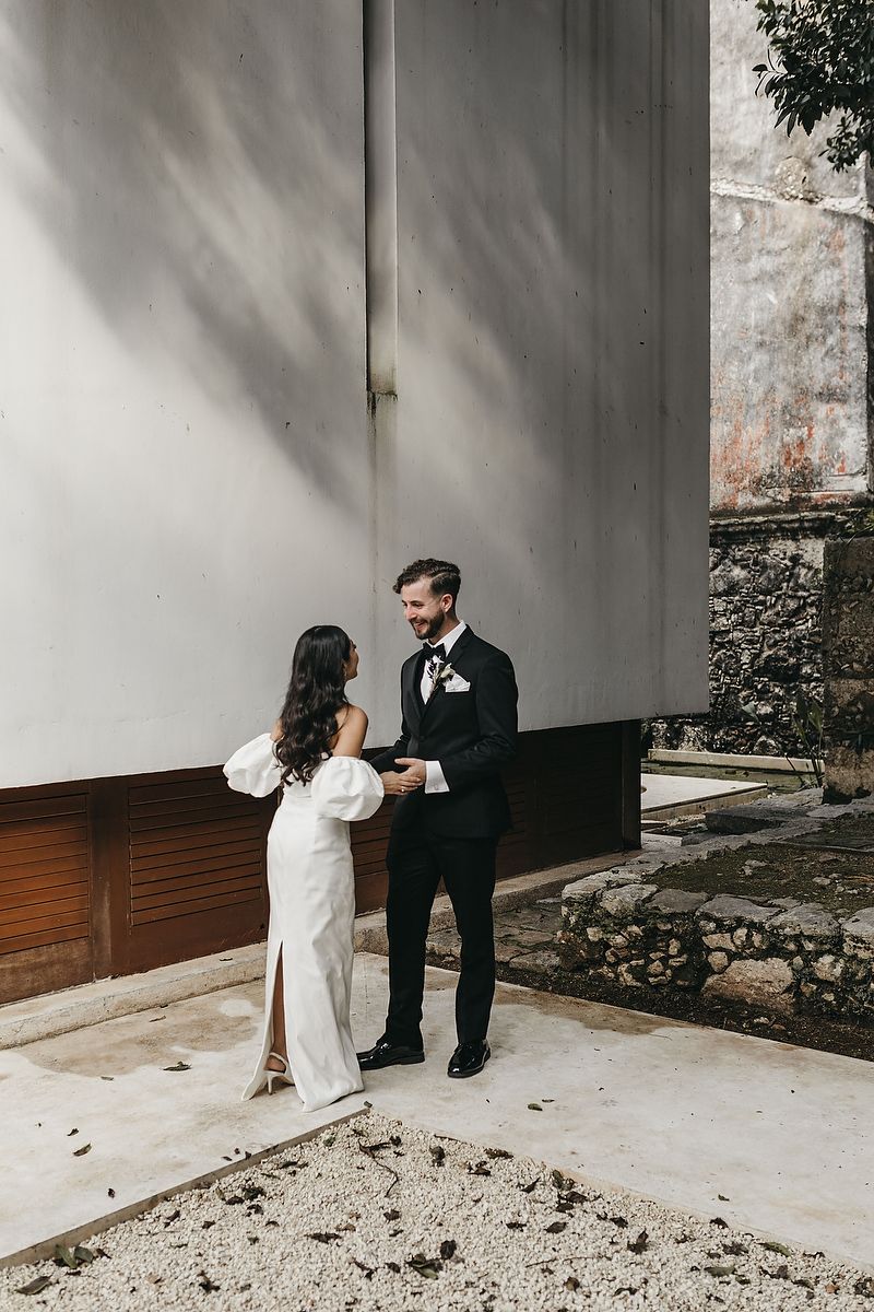 Smiling groom in black tuxedo holds bride's hands during an intimate first look moment at their wedding.