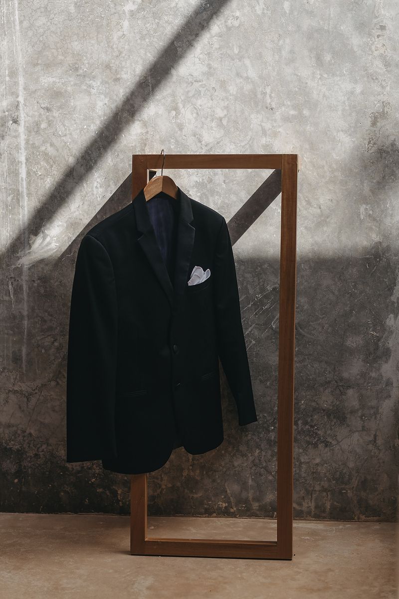 Dark navy blazer with white pocket square displayed on wooden clothing rack against concrete wall.