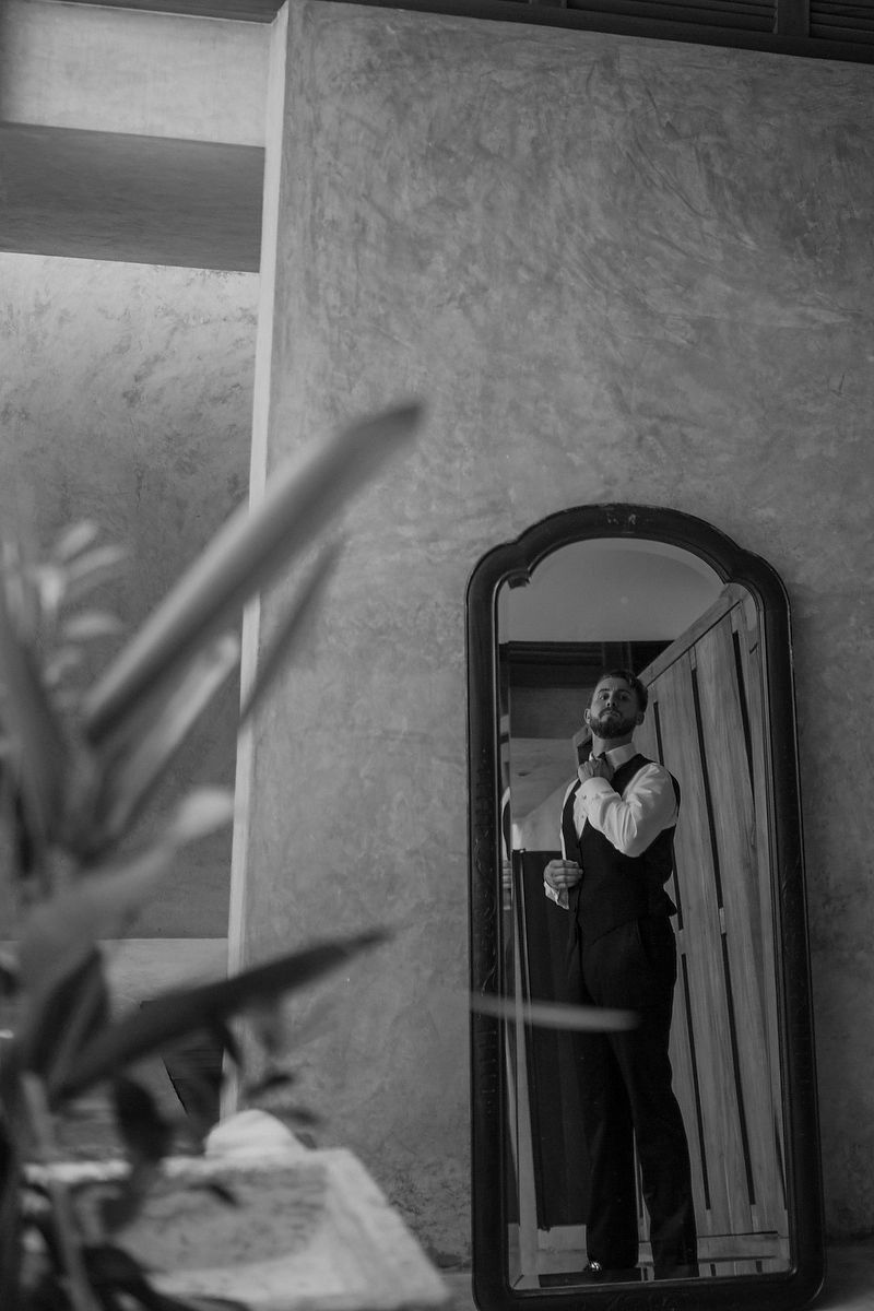 Groom adjusting tie in ornate floor mirror, black and white wedding preparation photo.