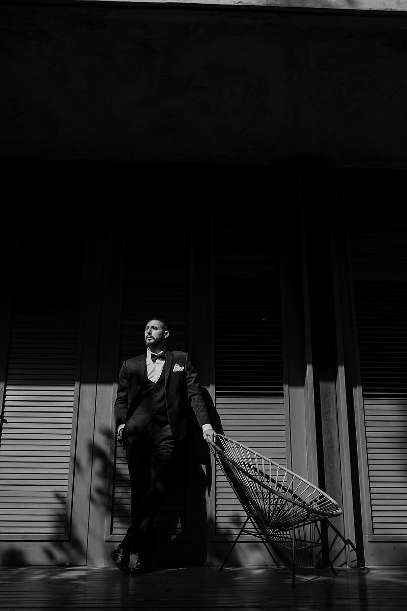 Elegant groom in tuxedo standing beside wire chair in dramatic black and white portrait.