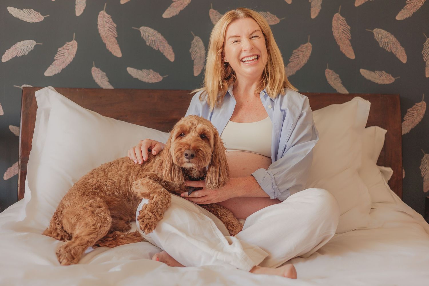 Golden doodle dog relaxing with owner on bed with feather pattern wallpaper.