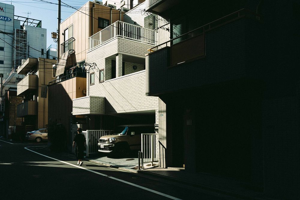A sunlit residential street in Japan with white buildings and balconies casting dramatic shadows.