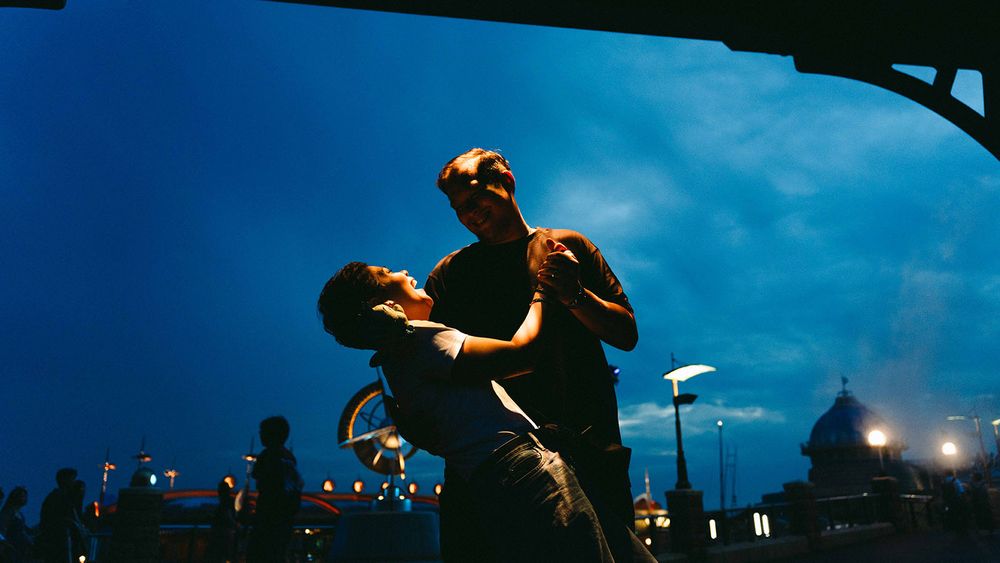 Silhouetted people jumping and dancing at dusk with colorful lights and a deep blue sky in the background.