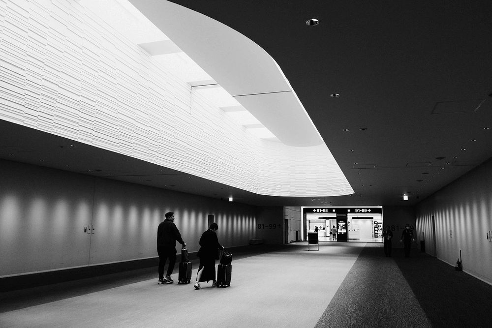 People traverse a sleek modern corridor with curved white walls and ambient lighting leading to a brightly lit exit doorway.