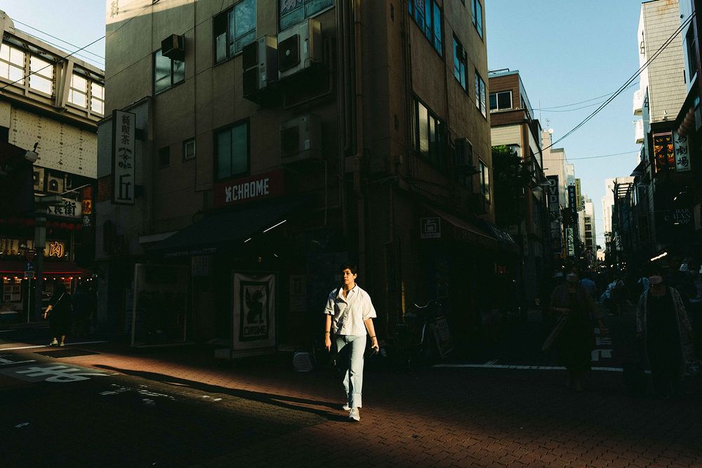 A person in a white outfit stands at a shadowy urban street corner in what appears to be an Asian city.