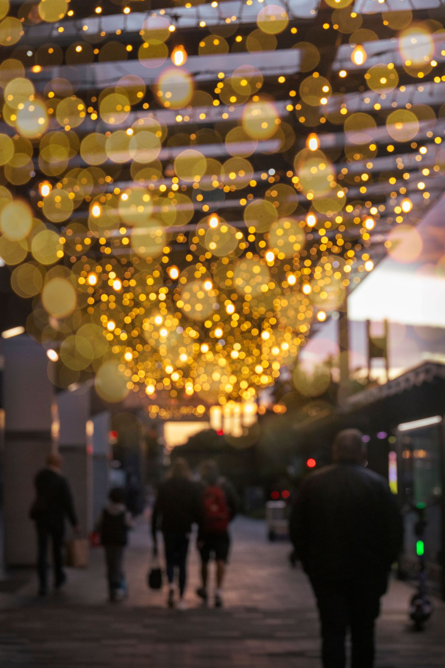 People walking beneath a canopy of golden glowing lanterns strung across a city street during evening twilight.