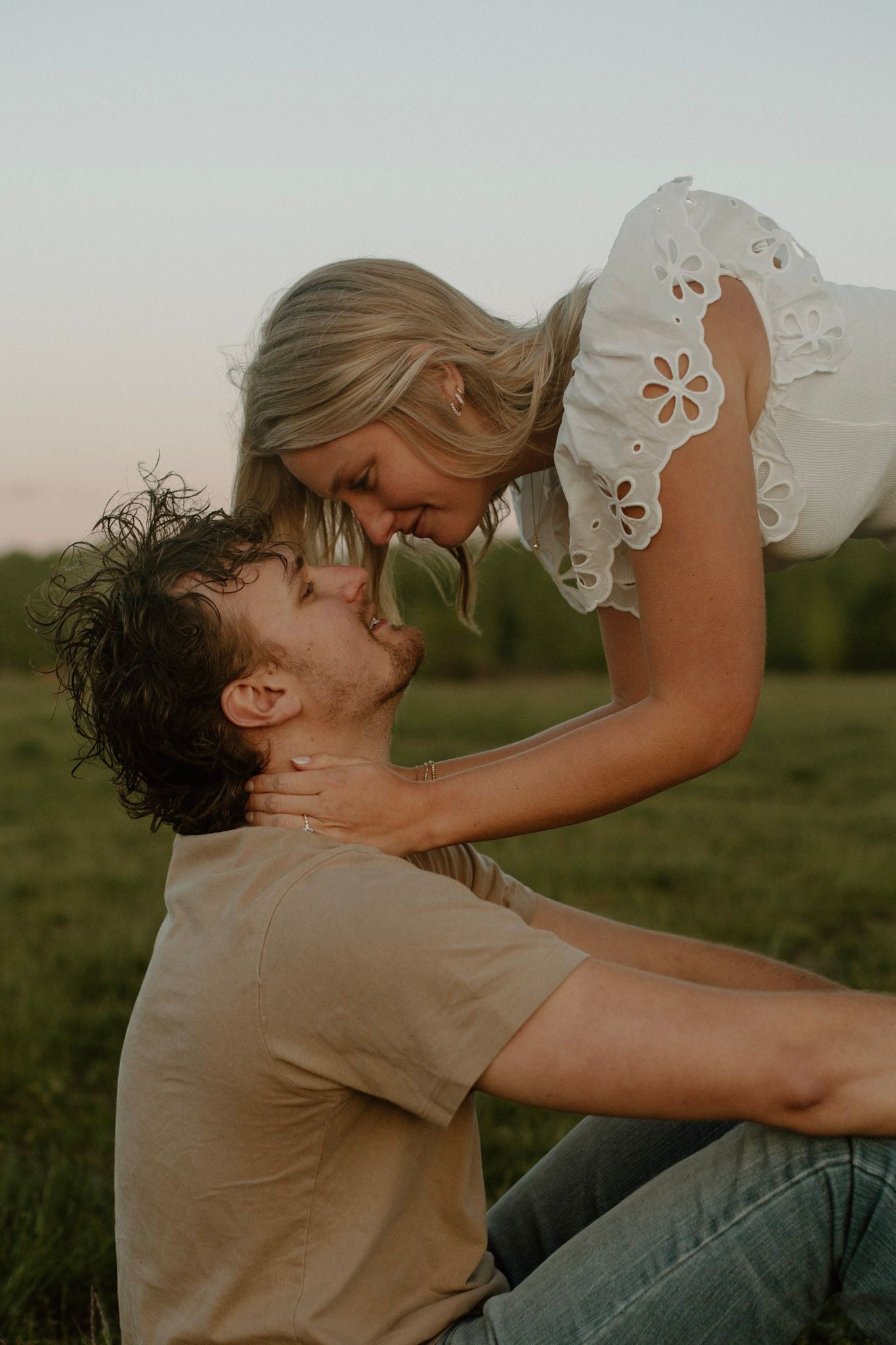 Couple in a field at golden hour engagement session. He is sitting with arms over his knees and she is leaning down towards him for a kiss.