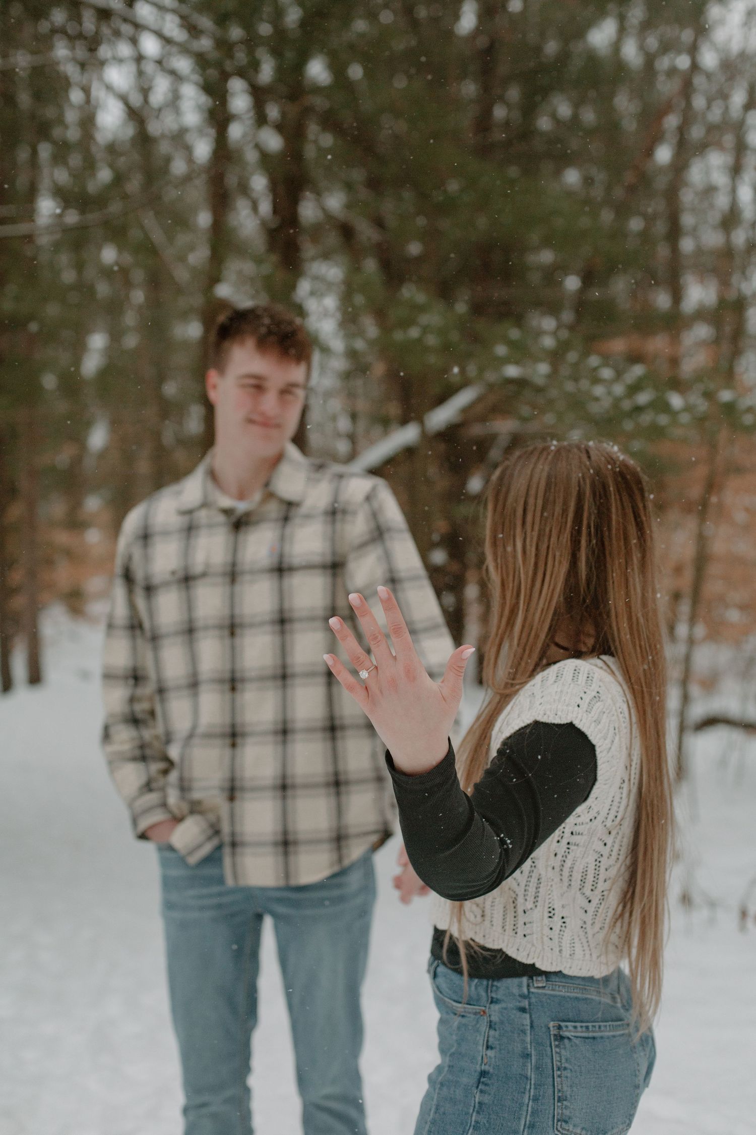 Couple during their winter engagement session in Holland, MI where she is showing off her ring while they look at each other.