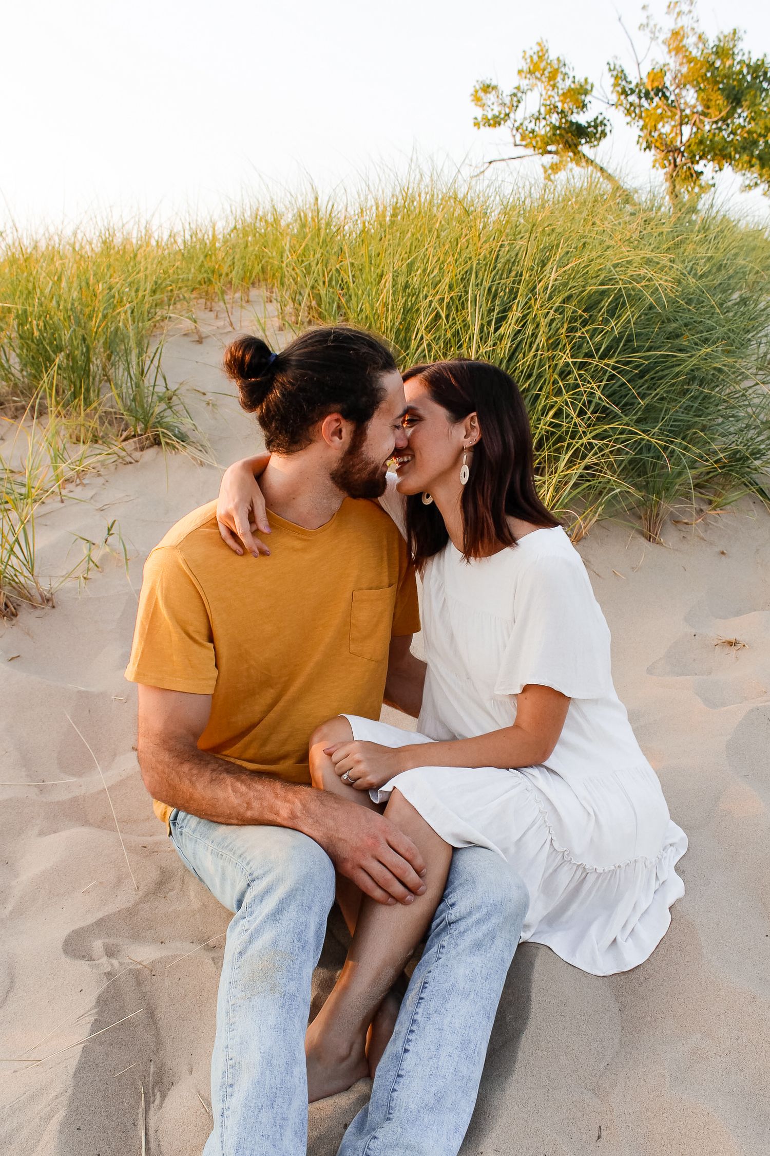 Couple sitting in the sand and embracing in front of dune grass in Holland, MI during the couple session.
