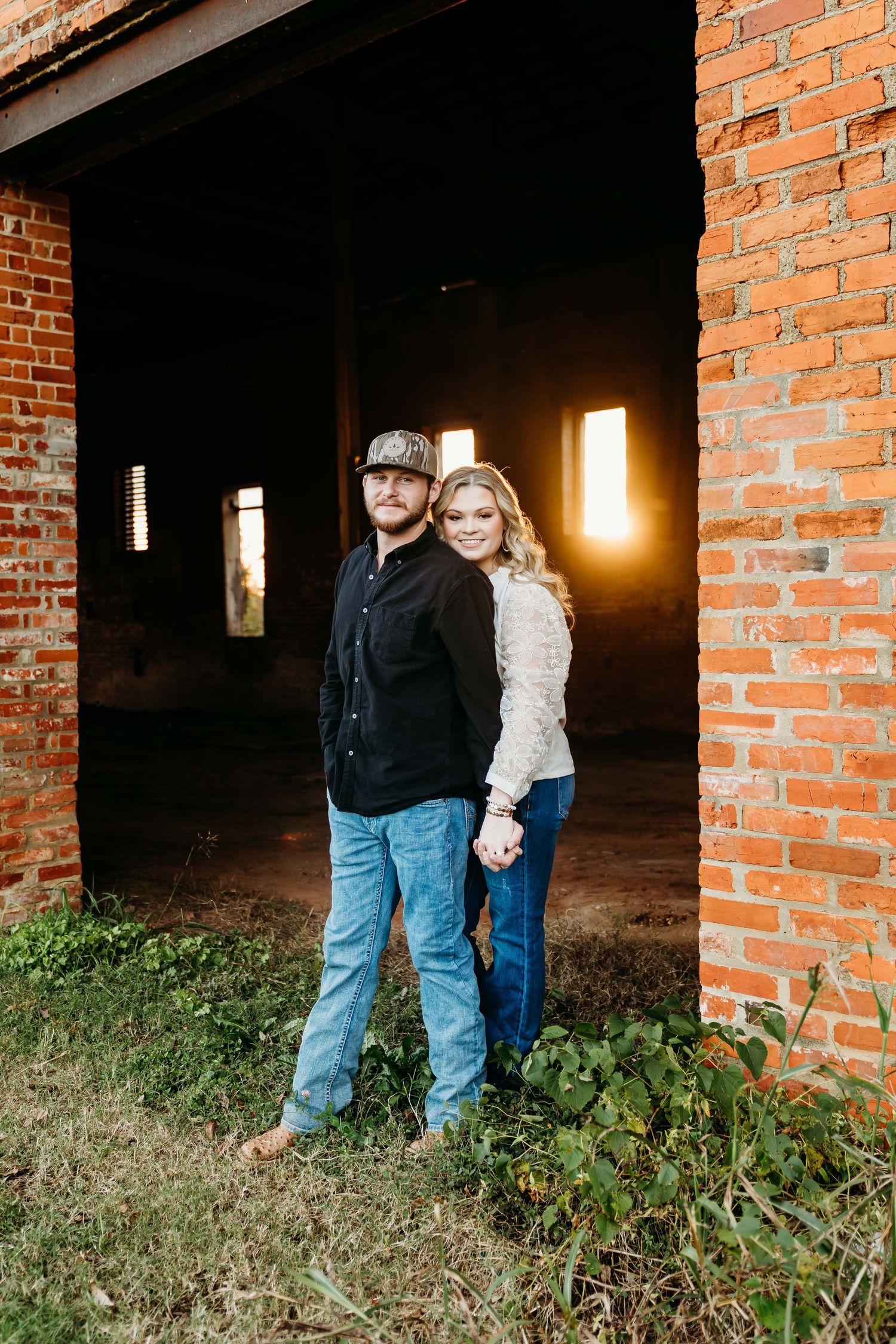 A couple stands together in front of a rustic brick doorway with warm natural lighting pouring in from the side.