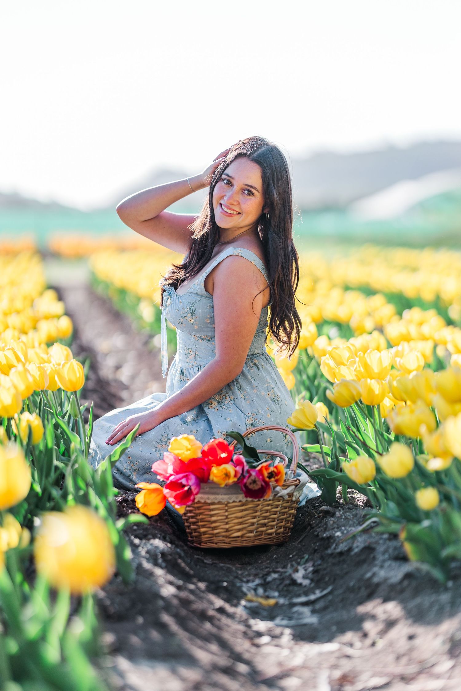 Woman in a sundress kneels in field of yellow tulips with colorful flowers in woven basket on sunny spring day at Underwood Family Farms.