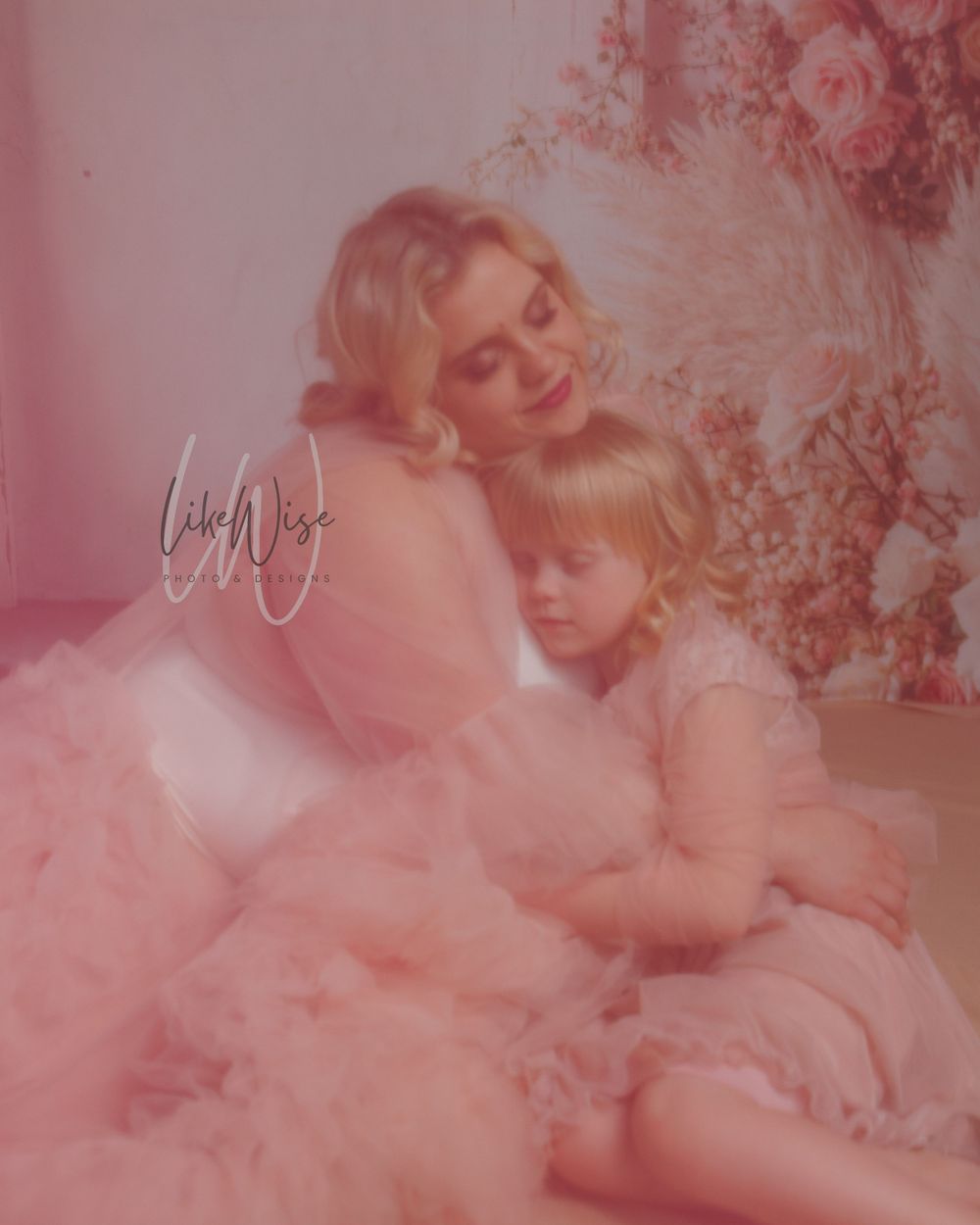 A dreamy pink-tinted photograph shows two figures in soft flowy gowns embracing amid floral decorations.